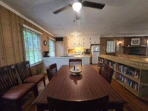 Rustic dining area with a long wooden table and chairs, wood-paneled walls, and a kitchen in the background.