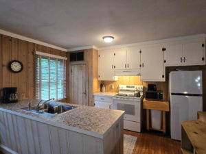 Kitchen with wood panel walls, white cabinets, and marble countertops; island sink in foreground and window with blinds on left edge.