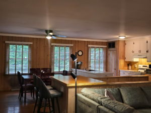 Cozy open-plan kitchen and dining area with wood-paneled walls, a ceiling fan, and large windows with blinds.