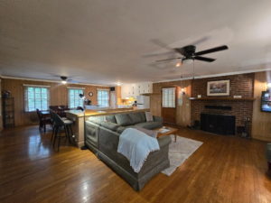 Open-plan living room with wood panel walls, a brick fireplace, and a ceiling fan; kitchen area visible in the background.