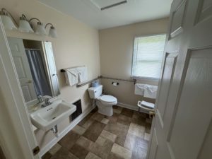 Small beige bathroom with sink, mirror, toilet, towel rack, and window blinds.
