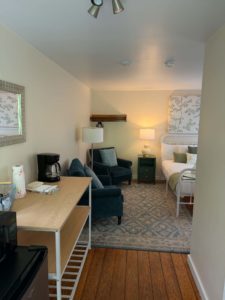 Hotel room with a desk and coffee station on the left, blue seating, and a bed at the far end with soft lighting and a patterned rug.
