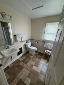 Small bathroom with sink, mirror, toilet, and towel bars; window with blinds, shower curtain to the left, tiled floor.