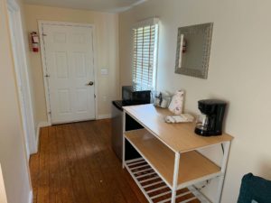 Narrow utility/kitchen corner with a white paneled door on the left, a mountedFire extinguisher, a window with blinds, and a desk holding a microwave and a coffee maker.