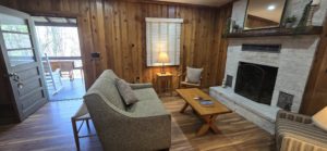 Cozy living room with wood-paneled walls, gray sofa, and a stone fireplace facing a wooden coffee table and chair. A lamp glows by a window with blinds; outside deck is visible through a door.
